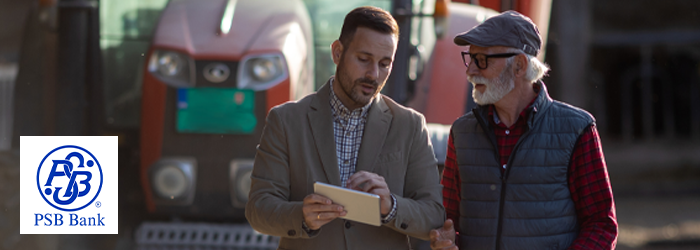 Farmer and banker looking at information on tablet while walking outside past farm equipment