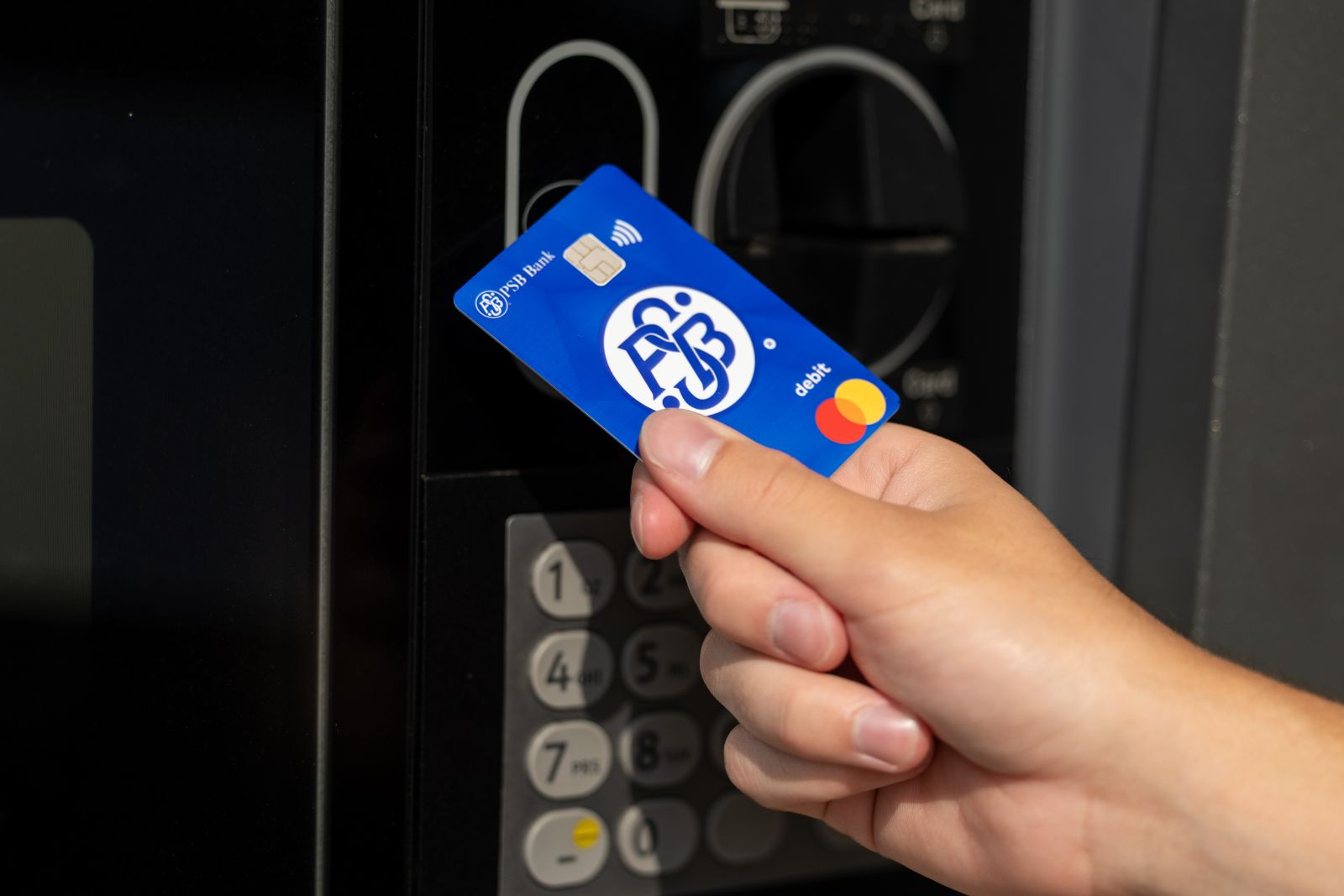 A hand holds a blue PSB Bank debit card in front of an ATM keypad and card reader.