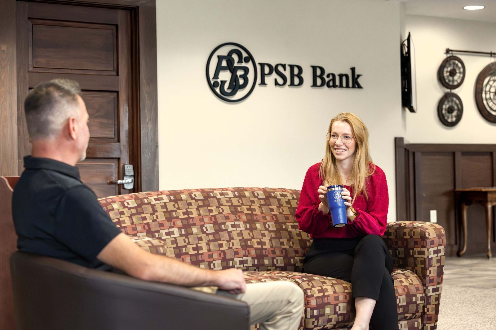 Two people having a conversation in the seating area of a PSB Bank branch, with the PSB Bank logo displayed on the wall behind them.