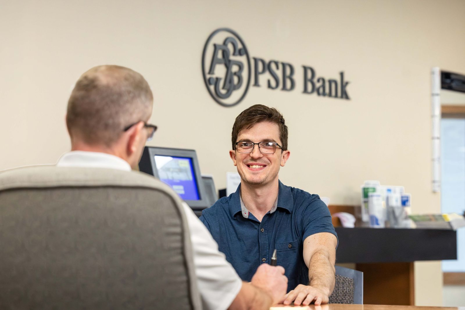 A PSB Bank employee assisting a customer at a desk inside a branch, with the PSB Bank logo displayed on the wall behind them.