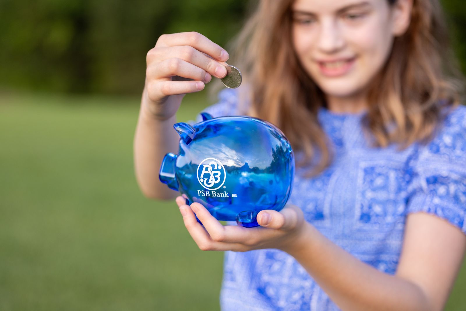 Young girl placing a coin into a blue PSB Bank piggy bank outdoors.
